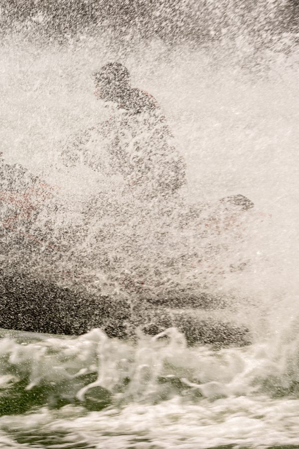 A jet ski making progress through a large amount of spray across the wake of a boat that is not shown in the image. The rider and jet ski are largely obscured by the spray and white foam of the wake.
